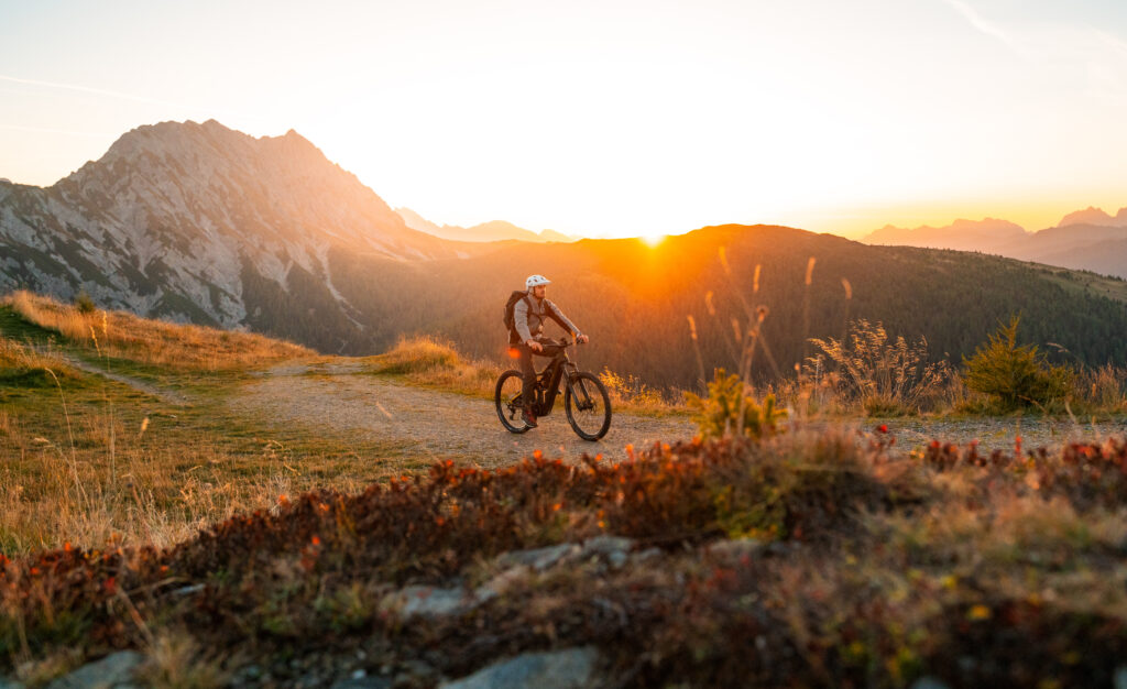 ebike-golzentipp-bergwiesentrail-herbst23-c-peter-maier-3_TVB Osttirol_Peter Maier_Obertilliach Mountainbiker bei Sonnenaufgang auf Alpenpfad mit Bergen im Hintergrund – Abenteuer und Outdoor-Aktivitäten in der Natur