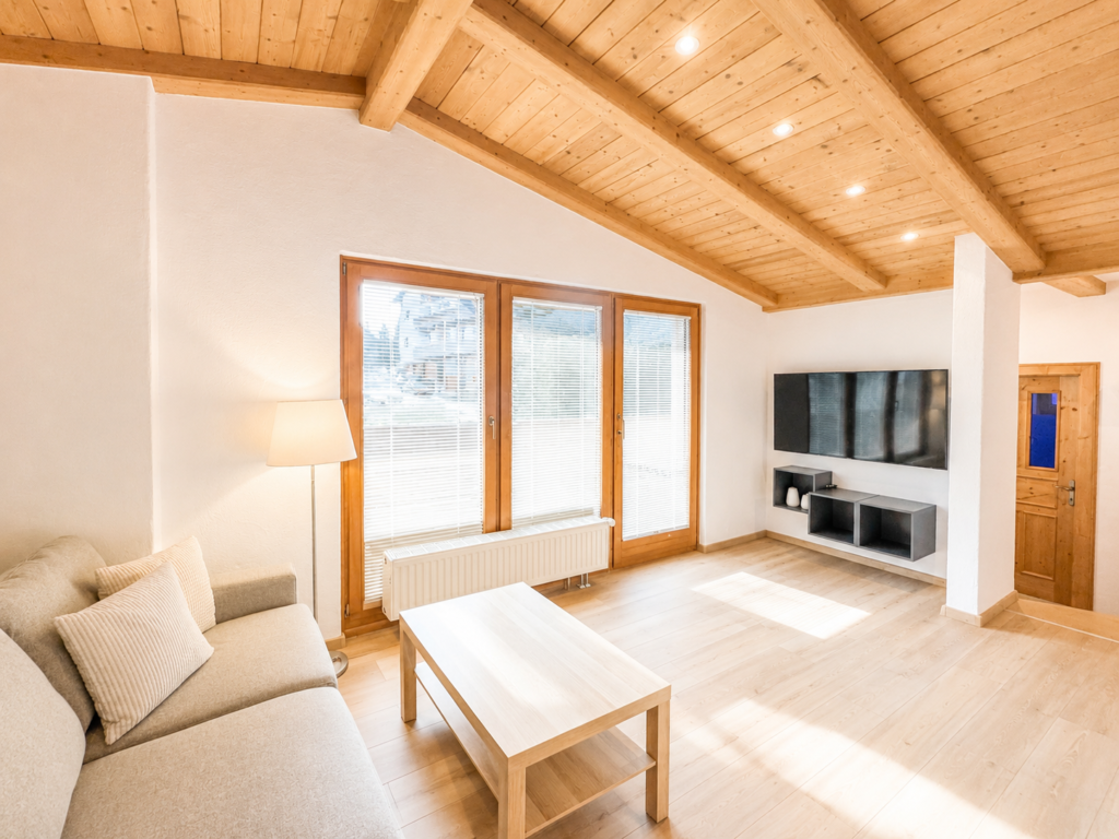Gemütliches Apartment mit Holzausstattung und modernem Wohnbereich Helles, modernes Wohnzimmer eines Apartments mit Holzdecke, Panoramafenstern, beigem Sofa, Holzcouchtisch und Flachbildfernseher auf weißer Wand