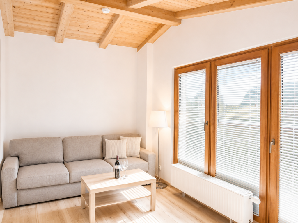 Komfortables Wohnzimmer mit Terrasse - Das Auss.Zeit Apartments Helles, modernes Wohnzimmer eines Apartments mit grauem Sofa, Holzbalkendecke, Holzterrasse mit großen Fenstern und Jalousien - Das Auss.Zeit Apartmenthaus