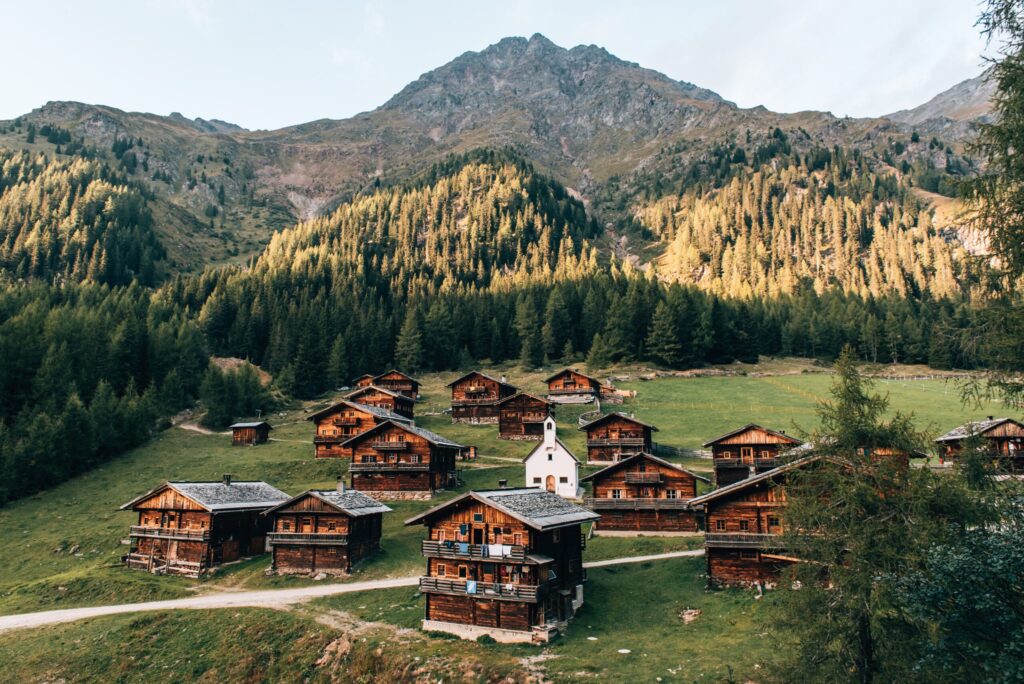 Oberstaller Alm_TVB Osttirol_Romeo Felsenreich_Innervillgraten Alpines Bergdorf mit traditionellen Holzhäusern und weißer Kapelle in einem Tal, umgeben von Lärchenwäldern und Bergen - Kulisse für Ruheappartements in den Alpen
