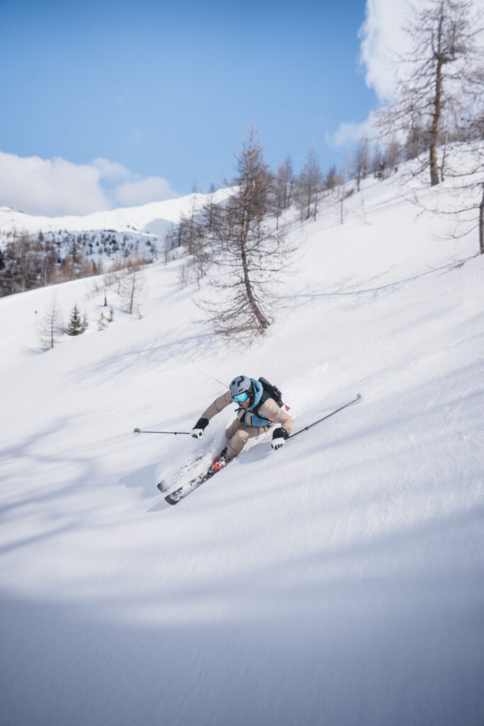 img 0073 Skifahrer in Carving-Position auf verschneiter Piste mit Lärchenbäumen und Berglandschaft im Hintergrund