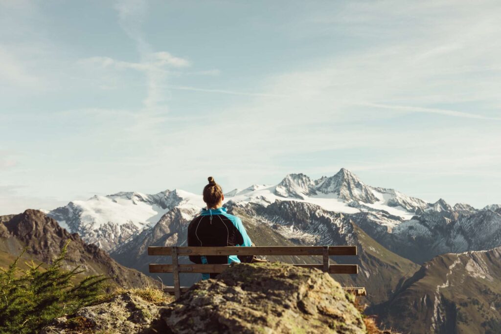 2000×1334 (1) Person sitzt auf Bank mit Blick auf schneebedeckte Berge in alpiner Landschaft